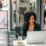 Ethnic young woman using laptop to automate digital ads while having tasty beverage in modern street cafe