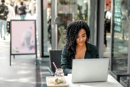 Ethnic young woman using laptop to automate digital ads while having tasty beverage in modern street cafe