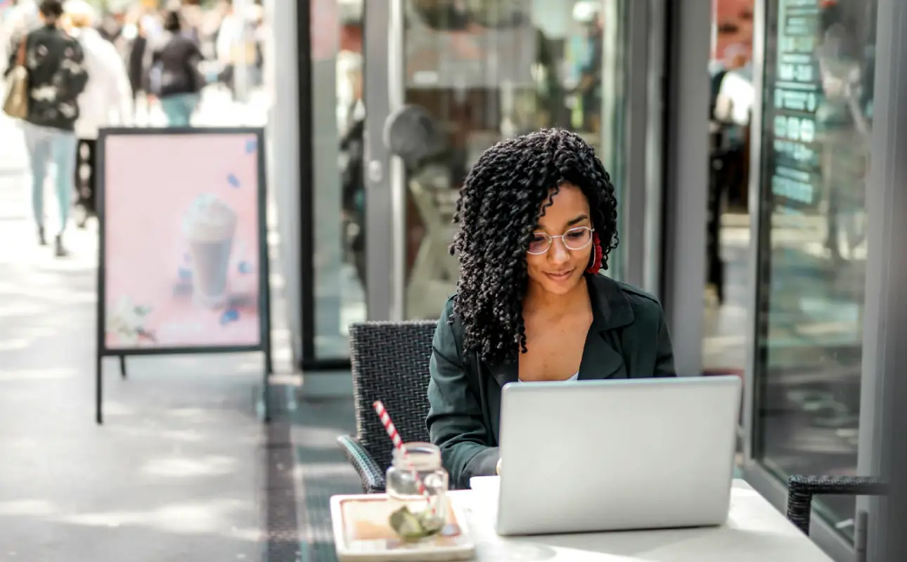 Ethnic young woman using laptop to automate digital ads while having tasty beverage in modern street cafe